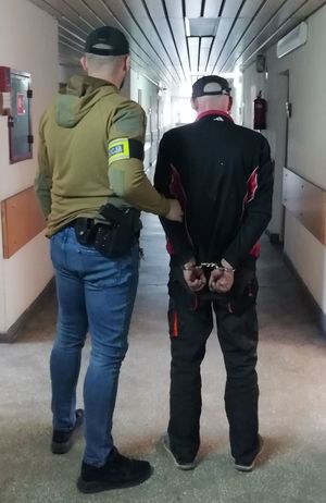 na korytarzu w budynku komendy nieumundurowany policjant z opaską na ramieniu  z napisem POLICJA prowadzi zatrzymanego który ma założone kajdanki na dłonie trzymane z tyłu