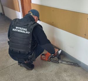 na korytarzy w budynku nieumundurowany policjant w kamizelce z napisem Wydział Kryminalny wykonuje oględziny pilarki spalinowej