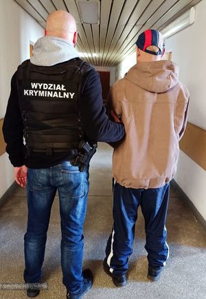 Na korytarzu komendy policjant ubrany w kamizelkę służbową prowadzi zatrzymanego.