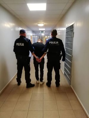 Obraz przedstawia dwóch funkcjonariuszy stojących na korytarzu Pomieszczenia dla Osób Zatrzymanych, gdzie trzymają mężczyznę które ma założone kajdanki