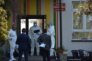 Na obrazku widocznych jest pięć osób, stojących przy wejściu do budynku. Trzy z nich ubrane są w białe kombinezony, kolejne dwie w marynarkach i spodniach.