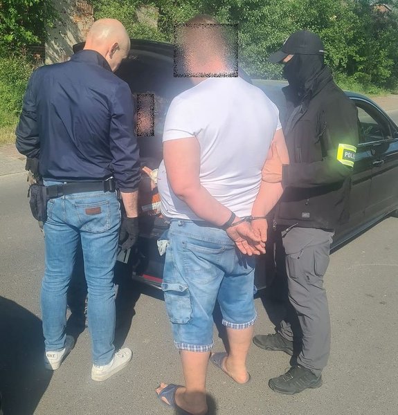 policjant przeszukuje bagażnik samochodu obok samochodu dwóch policjantów oraz zatrzymany mężczyzna który ma założony kajdanki na ręce trzymane z tyłu