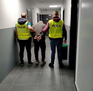 Na korytarzu komisariatu dwaj policjanci w kamizelkach odblaskowych z napisem Policja prowadza zatrzymanego.
