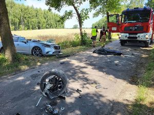 Miejsce wypadku drogowego, rozbity samochód srebrny który uderzył w  drzewo, porozrzucane elementy karoserii. Obok stoi  policjant i wóz strażacki oraz strażak.
