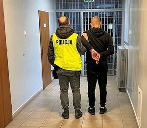 W areszcie policjant powadzi zatrzymanego który ma założone kajdanki na ręce trzymane z tyłu.