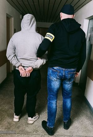 Na korytarzu komendy policjant z opaską na ramieniu prowadzi zatrzymanego który ma założone kajdanki na ręce trzymane z tyłu.