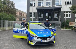 Na zdjęciu widoczny radiowóz policyjny zaparkowany  przed budynkiem, obok pojazdu stoją funkcjonariusze policji.