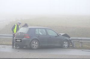 Dwaj umundurowani policjanci stoją przy samochodzie osobowym koloru grantowego, który uderzył w bariery ochronne przy drodze, samochód ma uszkodzony przód.