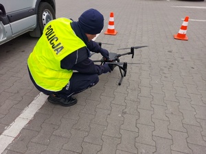 Policjant ubrany w kamizelkę odblaskową pochyla się nad dronem.