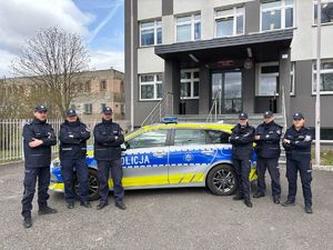 Przed budynkiem komendy stoi radiowóz a przed nim umundurowane policjantki i policjanci.