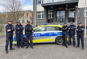 Przed budynkiem komendy na tle radiowozu stoją umundurowani policjanci.