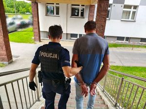 Umundurowany policjant po schodach prowadzi zatrzymanego mężczyznę, który ma założone kajdanki na ręce trzymane z tyłu.