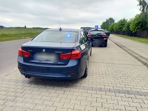 Na poboczu drogi stoi zaparkowany granatowy samochód, przed nim stoi auto z oznaczeniem na dachu literą &quot;L&quot;.