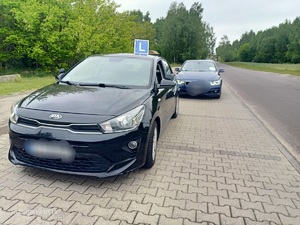 Na poboczu drogi stoi zaparkowane auto koloru czarnego z oznaczeniem na dachu literą &quot;L&quot;, za nim stoi granatowy samochód osobowy.