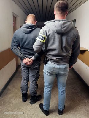 Na korytarzu komendy nieumundurowany policjant z opaską na ramieniu z napisem Policja prowadzi zatrzymanego mężczyznę.