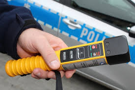 Na tle drzwi radiowozu widać trzymany w dłoni alkomat.