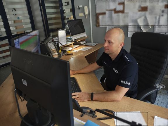Przy biurku przed monitorami komputerów siedzi umundurowany policjant.