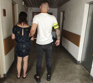 Na korytarzu komendy nieumundurowany policjant z opaską na ramieniu z napisem POLICJA prowadzi zatrzymaną kobietę.