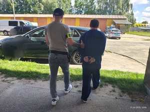 Nieumundurowany policjant z opaską na ramieniu prowadzi zatrzymanego do samochodu.