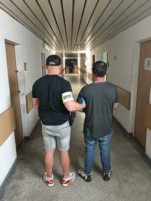 Nieumundurowany policjant z opaską na ramieniu prowadzi zatrzymanego po korytarzu komendy.