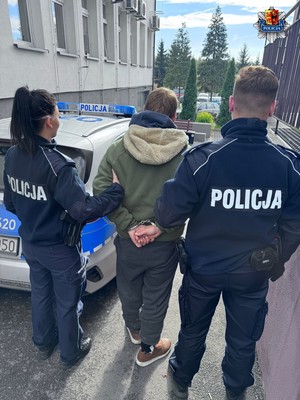 Przed budynkiem placówki oświatowej umundurowani policjanci prowadza zatrzymanego mężczyznę.