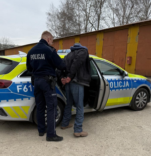 Do oznakowanego radiowozu umundurowany policjant wprowadza zatrzymanego mężczyznę, który ma założone kajdanki na ręce trzymane z tyłu.
