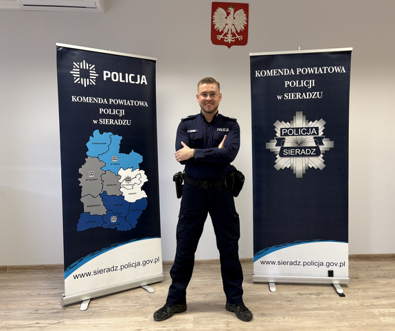 W pomieszczeniu służbowym na tle roll - up Komendy Powiatowej Policji w Sieradzu stoi umundurowany policjant.