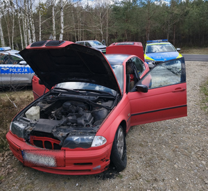 W kompleksie leśnym stoi samochód koloru czerwonego obok radiowozy.