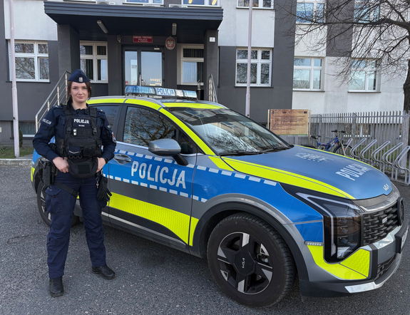 Przed budynkiem komendy na tle oznakowywanego radiowozu stoi umundurowana policjantka.