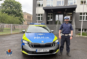 Przed budynkiem komendy przed oznakowanym radiowozem stoi umundurowany policjant.