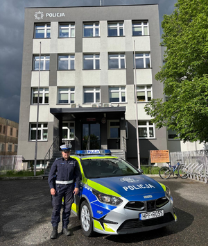 Przed budynkiem komendy przed oznakowanym radiowozem stoi umundurowana policjantka.