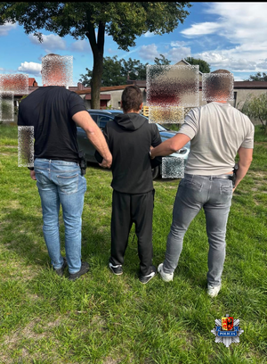 Przed posesją nieumundurowani policjanci prowadza zatrzymanego mężczyznę.