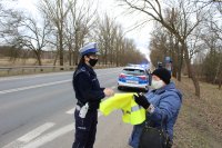 Na pierwszym planie umundurowana policjantka Wydziału Ruchu Drogowego wręcza kobiecie kamizelkę odblaskową, w tle radiowóz oznakowany oraz policjant WRD.