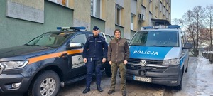 Umundurowany policjant stoi obok umundurowanego strażnika leśnego na tle radiowozu policyjnego typu bus oraz terenowego samochodu służbowego straży leśnej