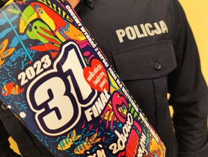 Na zdjęciu na tle policyjnego munduru i napisu &quot;Policja&quot; na pierwszym planie znajduje się kolorowa puszka do kwestowania z serduszkiem i logo Wielkiej Orkiestry Świątecznej Pomocy