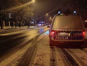 Zdjęcie zrobione wieczorem, na pierwszym planie na oświetlonej ulicy przy chodniku widać tył zaparkowanego radiowozu policyjnego, w tle z przodu widać pojazdy oraz dużą grupę osób.