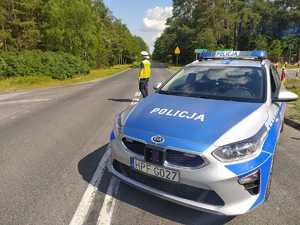 Policjantka Wydziału Ruchu Drogowego stoi na skrzyżowaniu i kieruje ruchem. Obok niej stoi radiowóz policyjny.