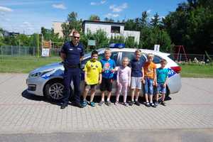 Dzieci wraz z policjantem pozują do grupowego zdjęcia. W tle widać radiowóz.