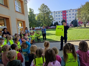 Policjant omawia konieczność noszenia elementów odblaskowych. Pokazuje dzieiom kamizelkę odblaskową.