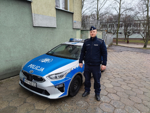 Umundurowany policjant stoi przed oznakowanym radiowozem.