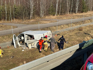 Poza drogą stoi uszkodzony bus. Ratownik, strażak oraz policjant prowadzą poszkodowanego.