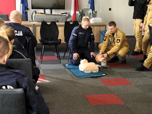 Policjanci ze strażakiem tłumaczą zebranym na szkoleniu policjantom jak odchylić ruchwę osoby poszkodowanej.