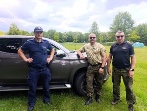 Policjant wraz z funkcjonariuszami straży rybackiej stoją przy samochodzie. W tle widać zbiornik wodny.