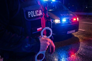Policjant trzyma kajdanki, w tle widać policyjny samochód.