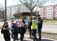 Policjanci w mundurach rozmawiają z dziećmi. Wręczają im elementy odblaskowe. W tle budynki i pojazdy.