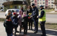 Policjanci w mundurach rozmawiają z dziećmi. Wręczają im elementy odblaskowe. W tle budynki i pojazdy.