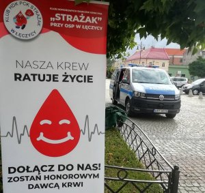 Zdjęcie przedstawia plakat Klubu HDK PCK „Strażak” przy OSP w Łęczycy z napisem „Nasza krew ratuje życie. Dołącz do nas zostań honorowym dawcą krwi”. Na drugim planie znajduje się oznakowany radiowóz policyjny.