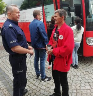 Zdjęcie przedstawia umundurowanego policjanta rozmawiającego z mężczyzną w czerwonym polarze z logo Klubu HDK PCK „Strażak” przy OSP w Łęczycy. Mężczyźni czekają w kolejce do krwiobusu.
