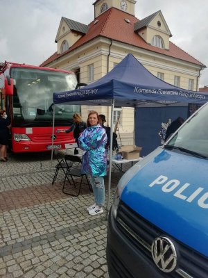 Zdjęcie przedstawia uśmiechniętą kobietę ubraną w błyszczącą fioletową kurtkę oraz spodnie jeansowe, stojącą przy stoisku promocyjnym KPP w Łęczycy pomiędzy krwiobusem a radiowozem policyjnym.