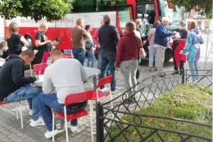 Na zdjęciu grupa osób czekająca w kolejce do mobilnego punktu poboru krwi przy ratuszu miejskim, dwóch mężczyzn siedzi przy stoliku i wypełnia formularze.
